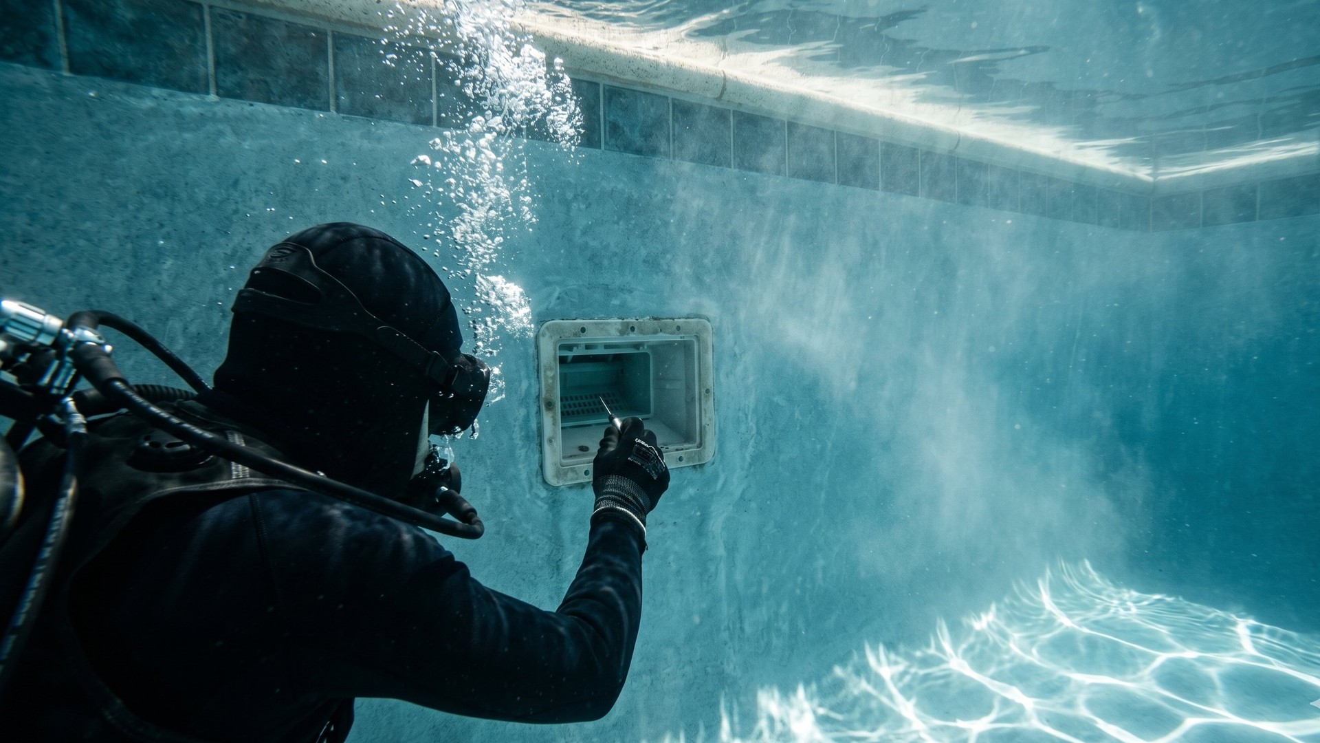 A diver in dark wetsuit and full air-supplied breathing apparatus inspecting a residential pool's skimmer fitting from underwater. Bubbles rise from the regulator; caustic light patterns ripple across the pool floor.